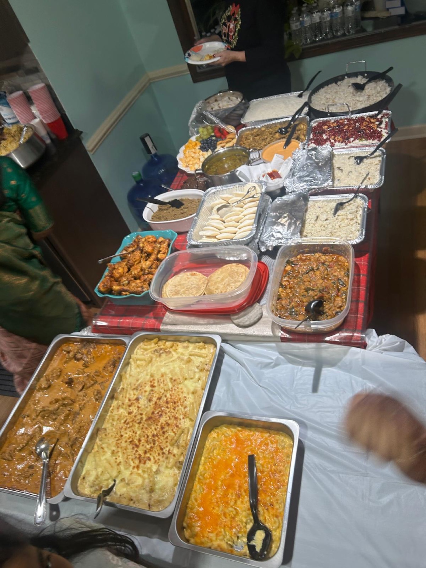 A long fellowship potluck table arranged with rice dishes, curries, baked dishes, breads, fruit, desserts, sauces, plates, and serving utensils